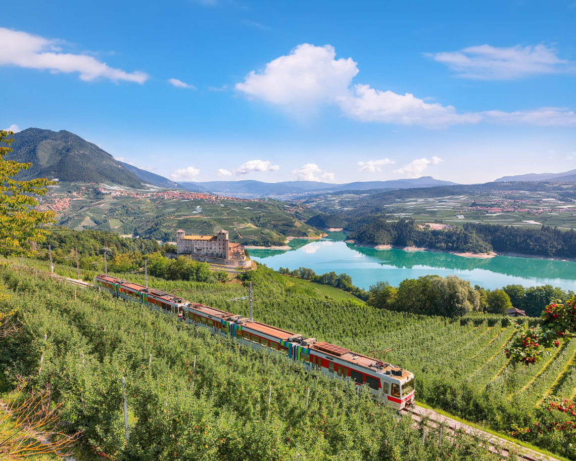 Regional train passing through lush apple orchards with Castel Thun castle and turquoise Lake Santa Giustina in the valley below, Val di Non, Trentino, Italy