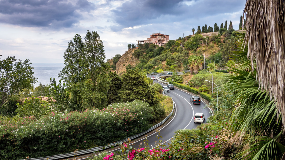 Winding road with cars curving through lush Mediterranean vegetation with bougainvillea, palm trees and a historic hilltop villa overlooking the Ionian Sea, Taormina, Sicily, Italy
