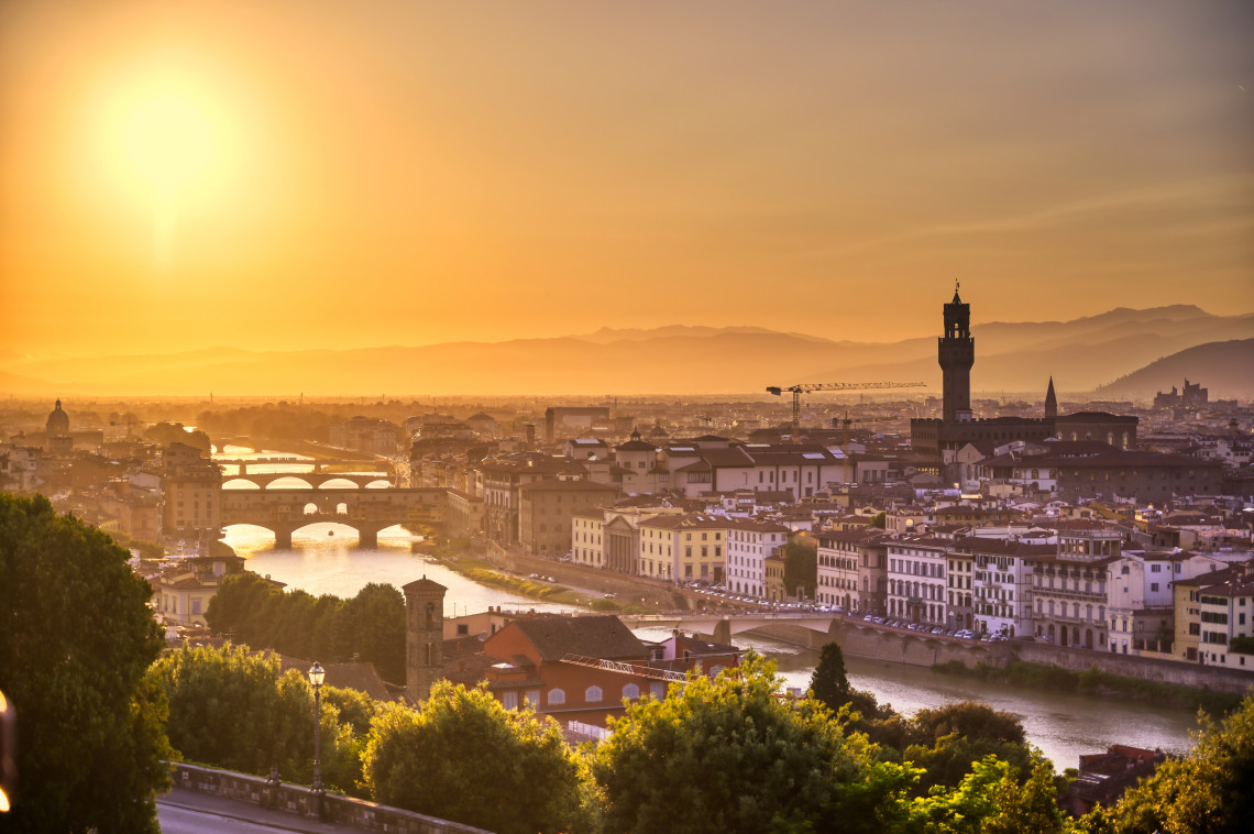 Panoramic sunset view over Florence, Italy, with the Arno River, historic bridges and Palazzo Vecchio rising above the city centre. The elevated perspective looks across Florence toward the surrounding Tuscan hills.