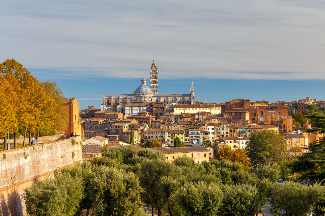 Siena Cathedral rises above the terracotta rooftops of Siena, Tuscany, Italy, with its striped marble exterior, dome and bell tower dominating the historic skyline. Olive trees and medieval buildings fill the foreground.