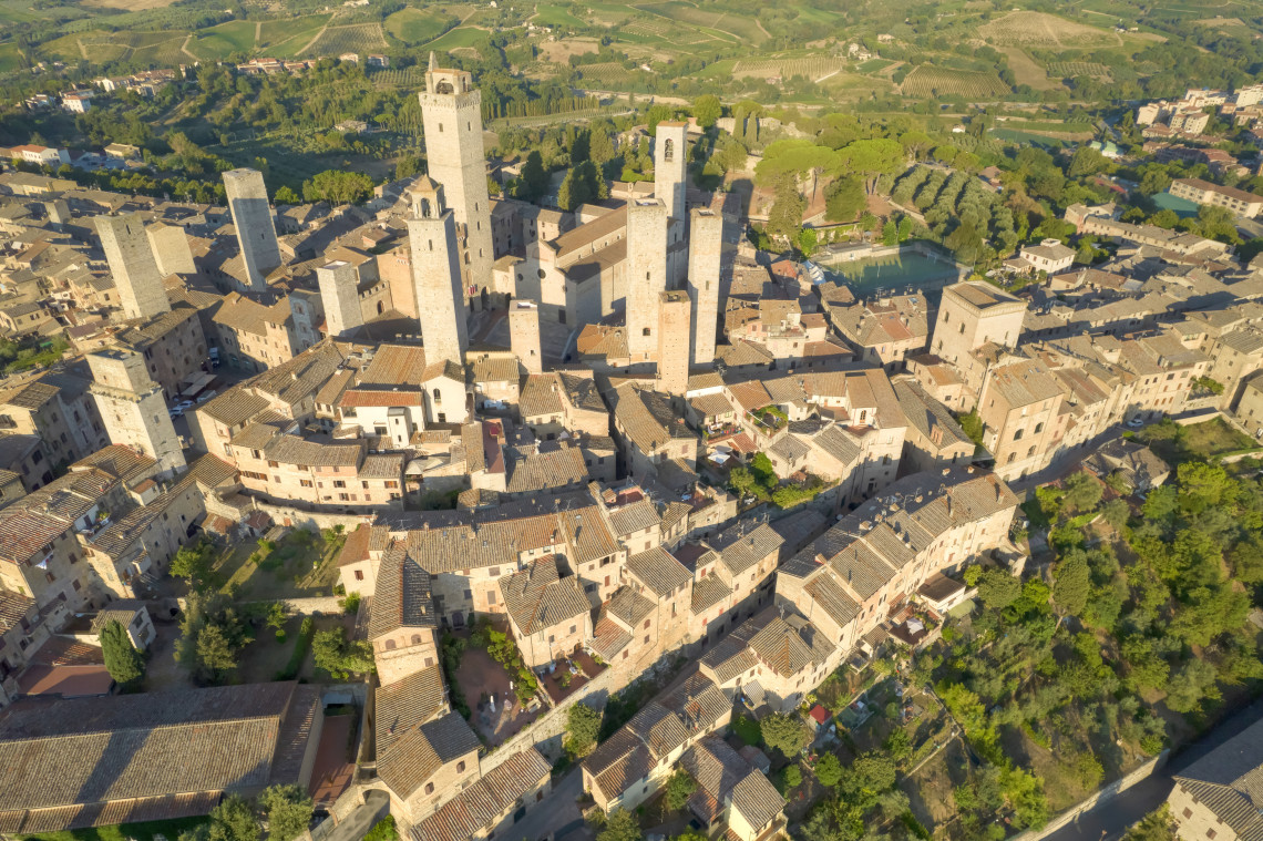 Aerial view of San Gimignano in Tuscany, Italy, showing medieval stone buildings and the tall towers that define the historic centre. Vineyards, farmland and green hills surround the fortified town.