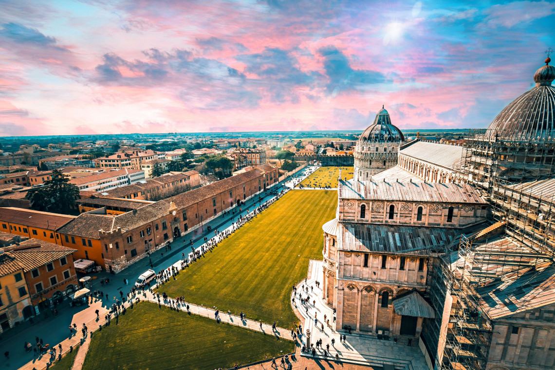 Elevated view across Piazza dei Miracoli in Pisa, Italy, showing Pisa Cathedral and its domed Baptistery beside the broad green lawn. The monumental religious complex forms part of Pisa&rsquo;s UNESCO-listed historic square.