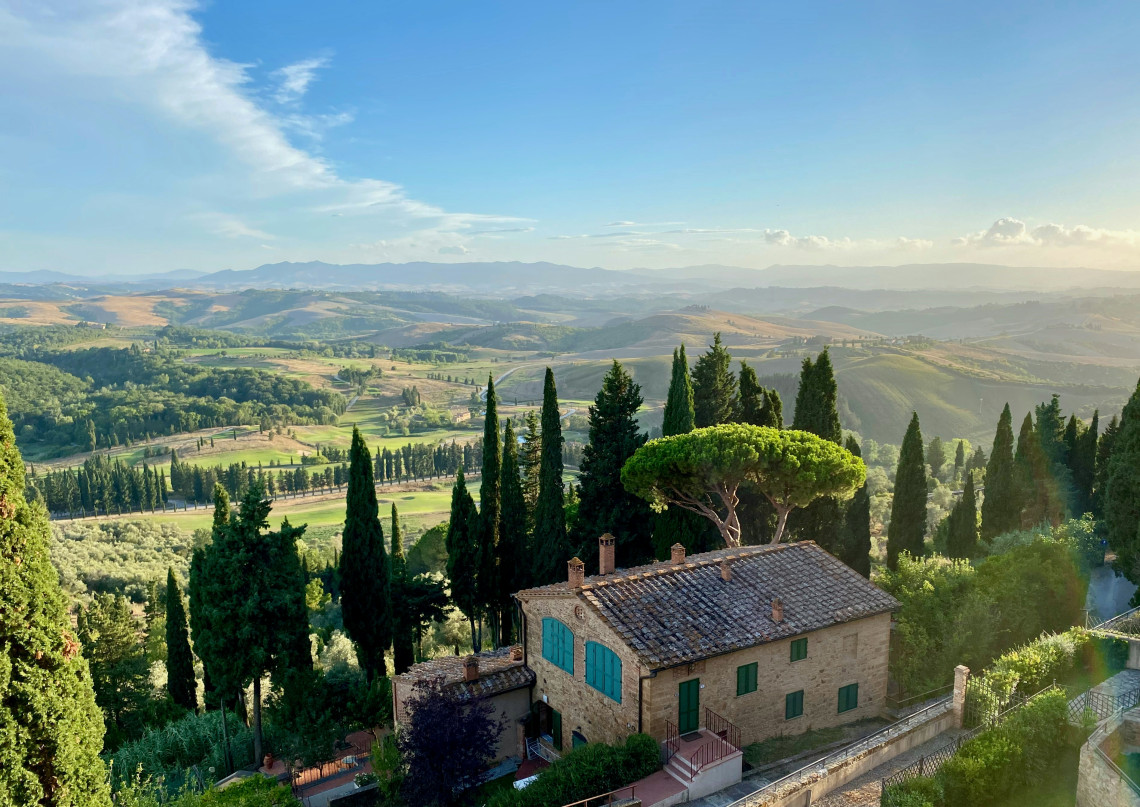 A stone farmhouse surrounded by cypress trees overlooks rolling hills and cultivated countryside in Tuscany, Italy. Olive groves, farmland and distant ridges extend across the rural landscape.