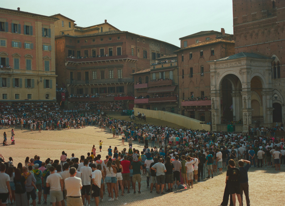 Crowds gather around Piazza del Campo in Siena, Italy, during the Palio di Siena, with the temporary racing track running around the medieval square. Historic palaces frame the packed public space during one of Siena&rsquo;s best-known traditions.