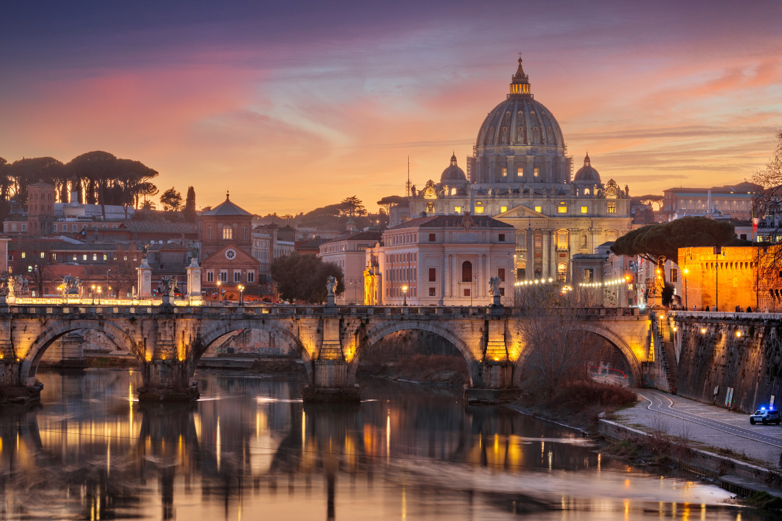 St. Peter&rsquo;s Basilica rises above Rome at sunset beyond the illuminated arches of Ponte Sant&rsquo;Angelo and the Tiber River. Warm evening lights reflect in the water beneath the Vatican skyline.