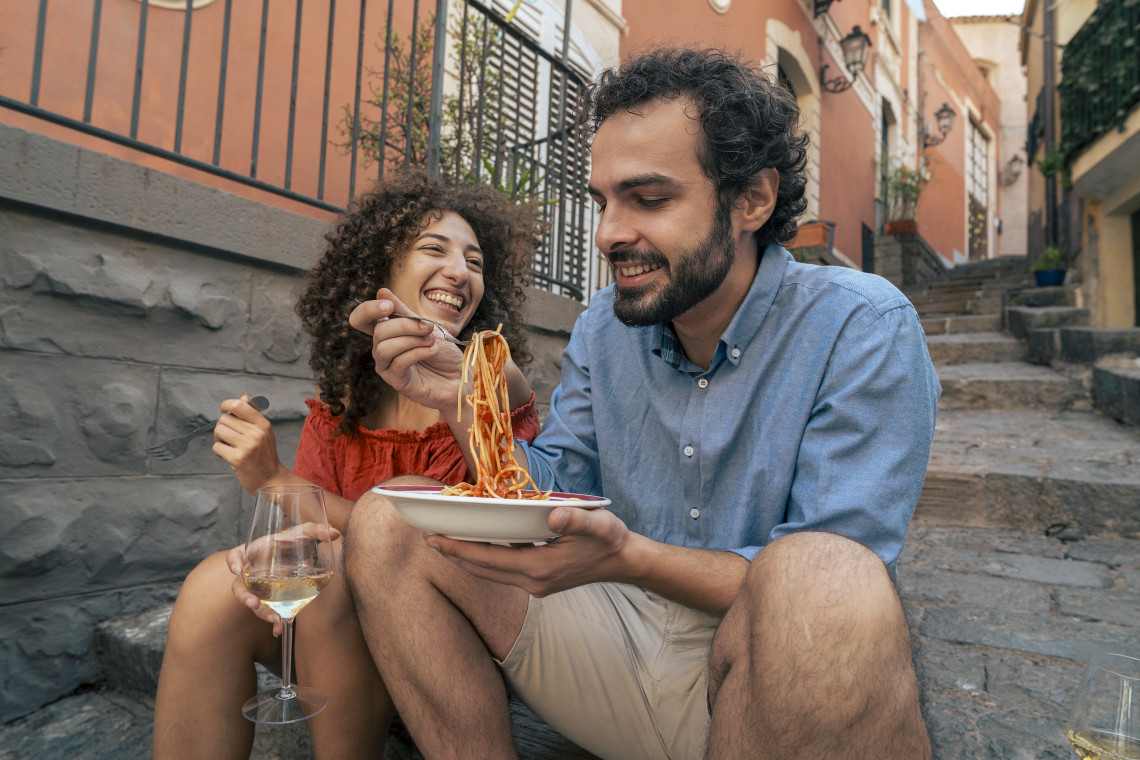 Couple laughing and sharing a plate of spaghetti with a glass of white wine while sitting on cobblestone steps in an Italian village alley