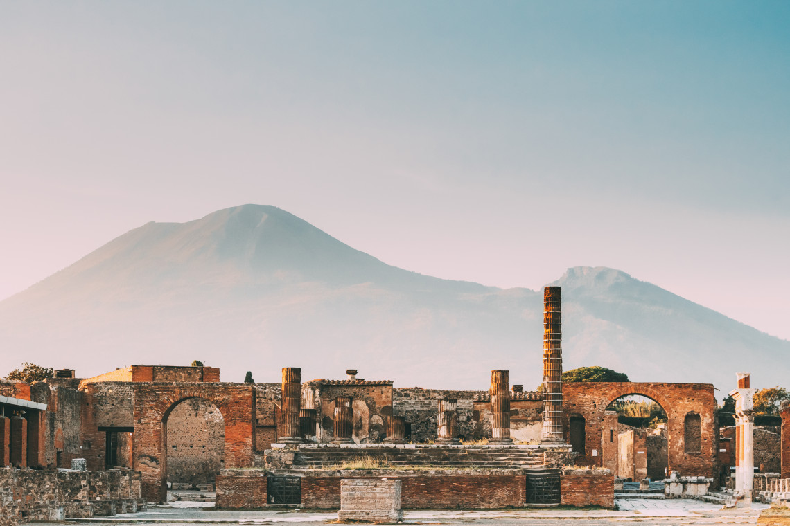 Ancient Roman ruins of Pompeii with brick arches and columns in the foreground and the silhouette of Mount Vesuvius looming in the misty background, Campania, Italy
