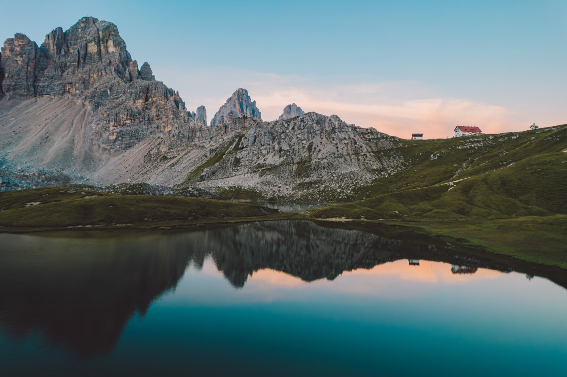 Mirror reflection of Tre Cime di Lavaredo rocky peaks and a red roofed mountain refuge in the still waters of Laghi dei Piani at sunset, Dolomites, South Tyrol, Italy