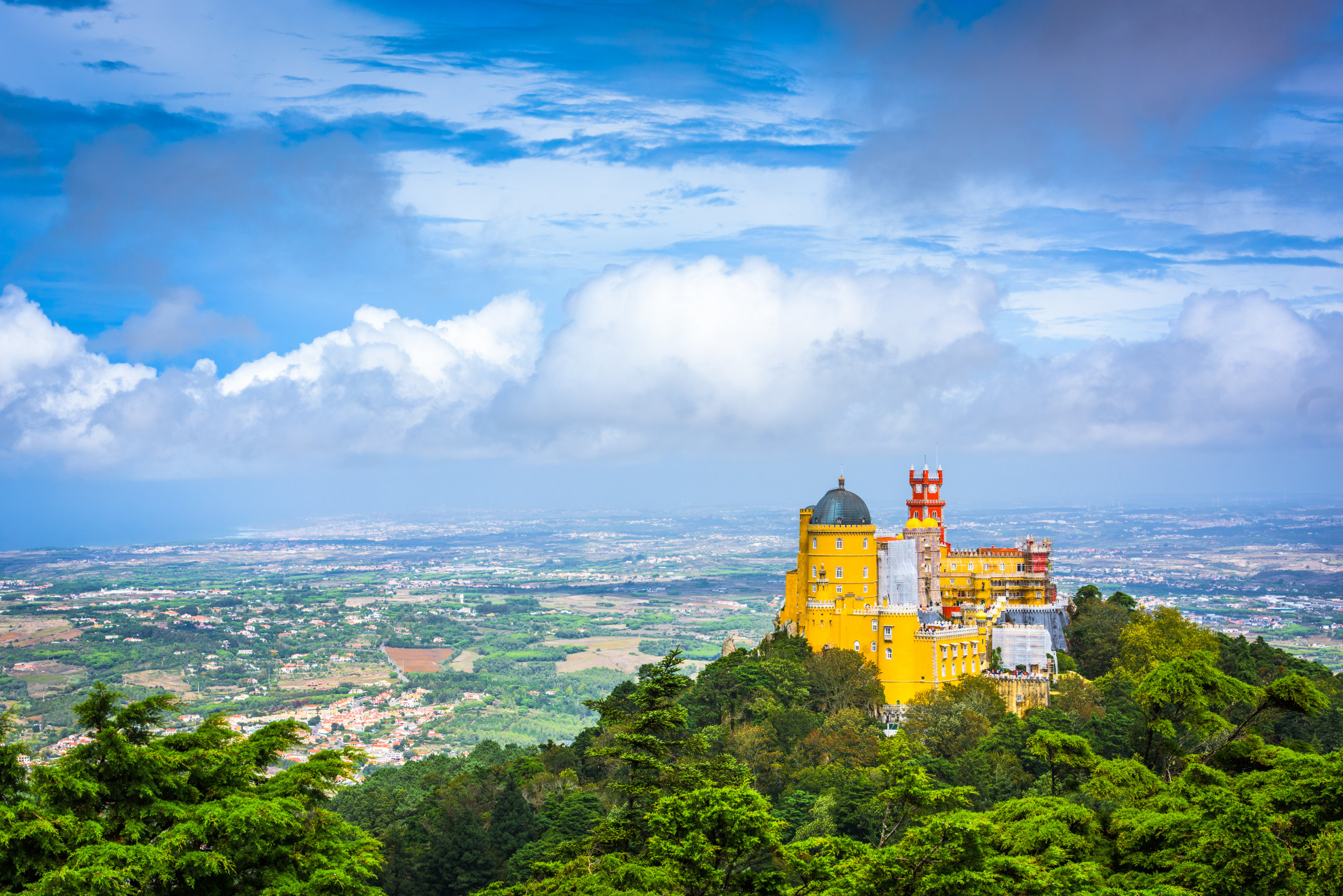 Colorful Pena National Palace sits on a hilltop above Sintra’s forests, with wide valley views under a cloud-filled sky.