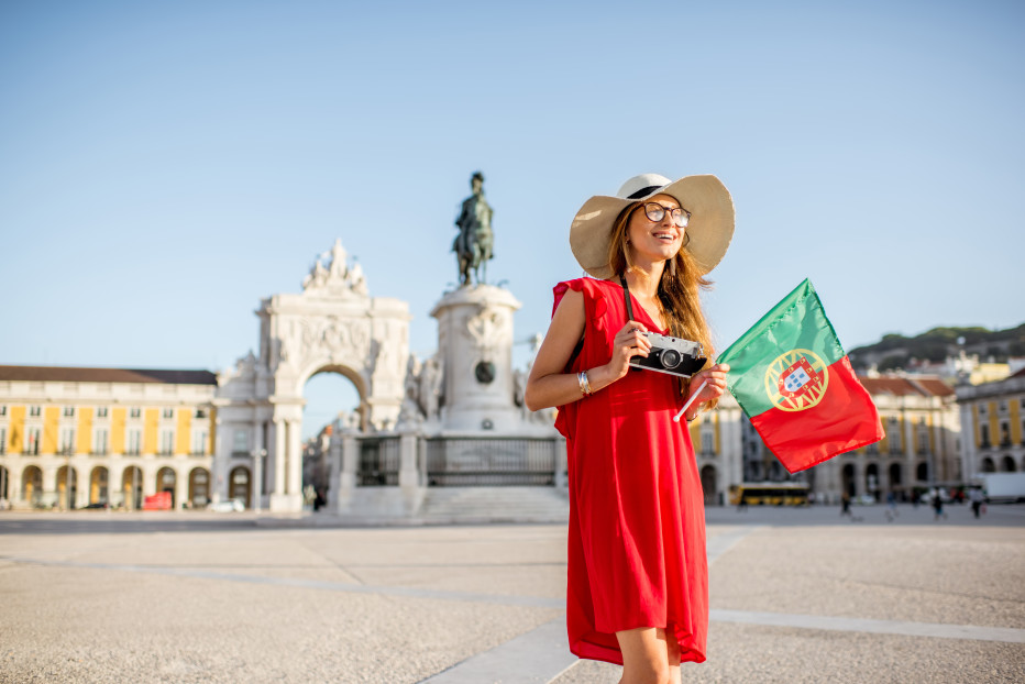 miling traveler in a red dress holds a Portuguese flag and camera at Lisbon&rsquo;s Pra&ccedil;a do Com&eacute;rcio, with the Arco da Rua Augusta and King Jos&eacute; I statue behind her on a sunny day.