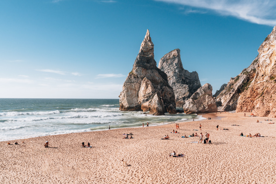 Beachgoers relax on the sand at Praia da Ursa near Sintra, with dramatic sea stacks, rugged cliffs, and Atlantic waves under a clear summer sky.