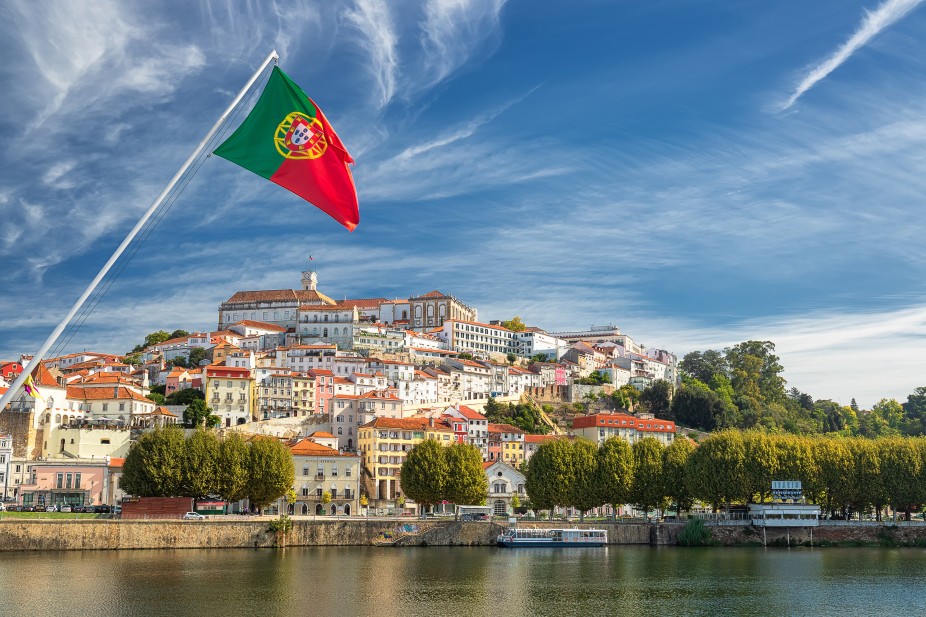 Portugal&rsquo;s flag waves above the Mondego River in Coimbra, with the hilltop old town and University of Coimbra skyline under a bright blue sky.