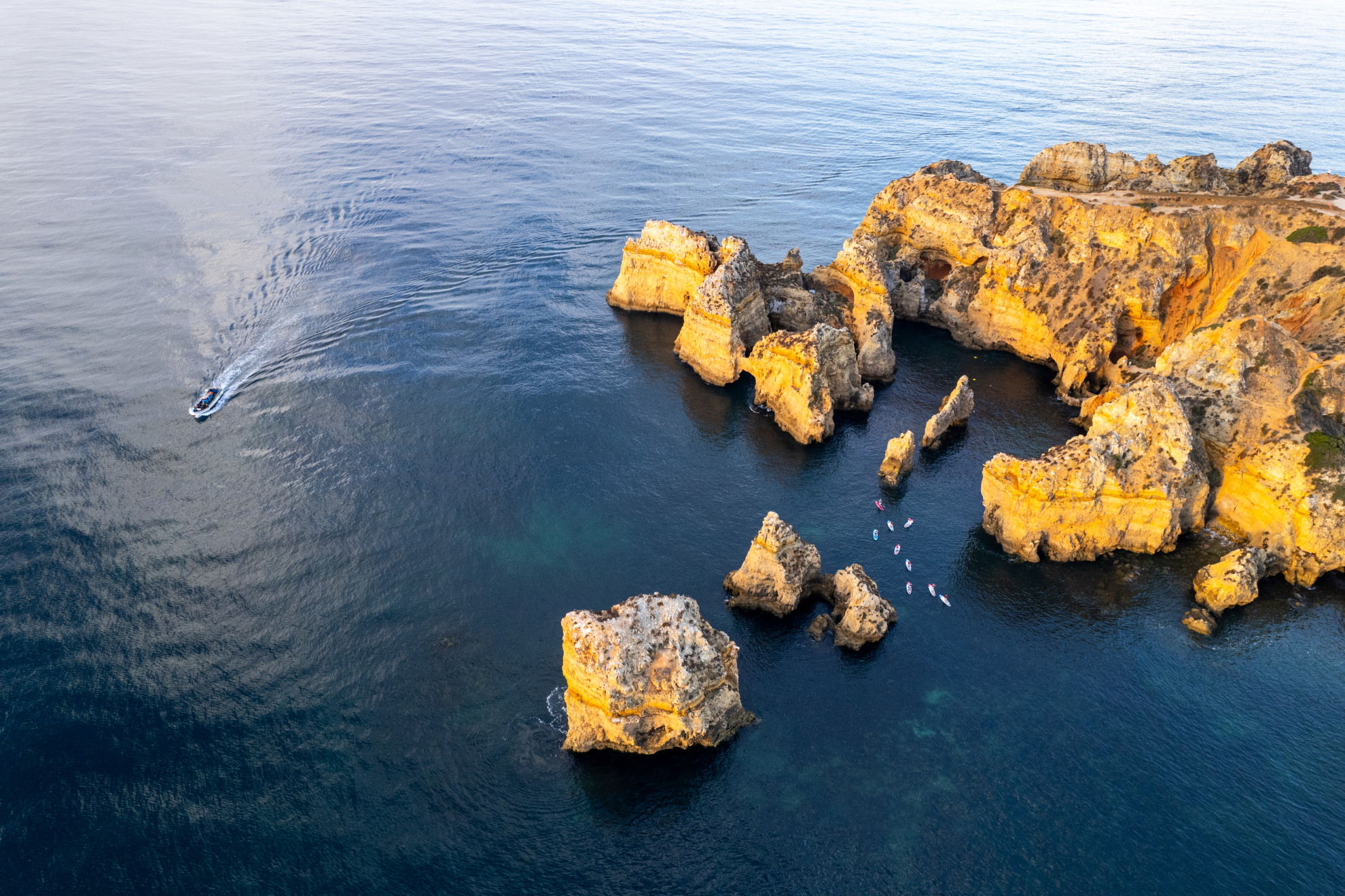Aerial view of Ponta da Piedade in Lagos, with golden limestone cliffs, natural arches and isolated sea stacks surrounded by deep blue Atlantic water as boats and kayaks explore the formations.