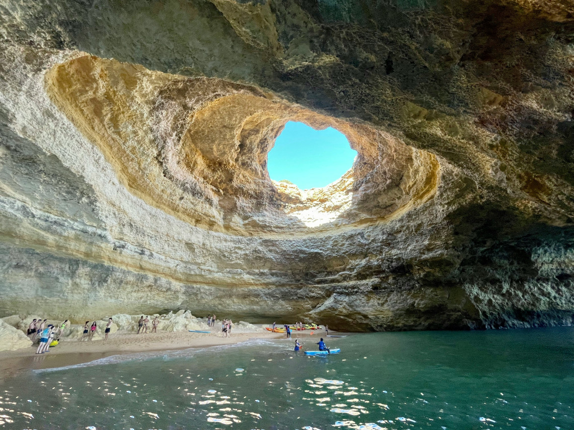 Interior view of Benagil Cave near Lagoa in the Algarve, Portugal, with sunlight entering through the circular opening in its limestone ceiling. Visitors and kayaks gather on the sandy shore beside the turquoise water.