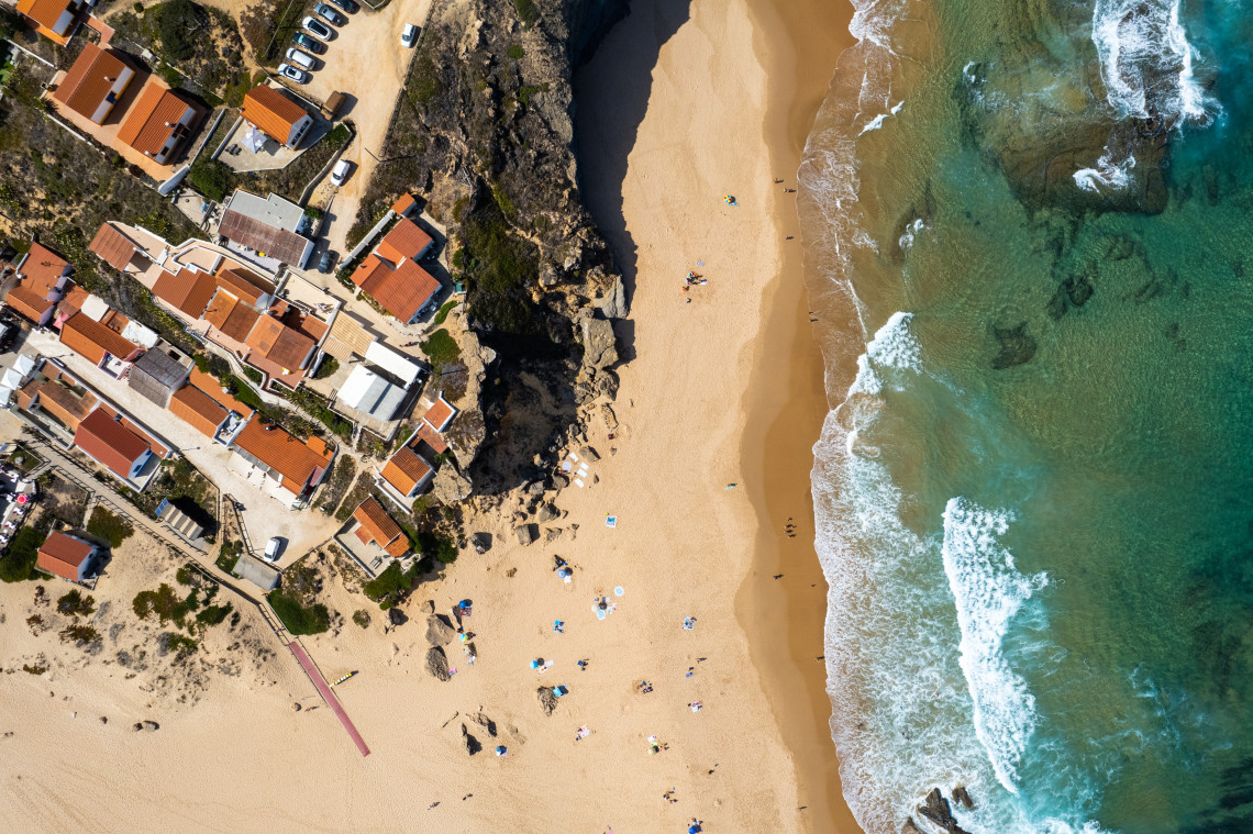Top-down aerial view of Praia da Gal&eacute; in Albufeira, Portugal, showing golden sand, clear coastal water and rock formations beside terracotta-roofed buildings. A beach access path leads from the residential area to the shoreline.