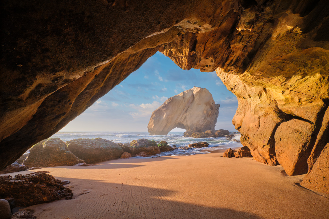 View from a coastal cave toward the natural Arco de Albandeira in Lagoa, with golden limestone formations, a sandy beach and Atlantic waves illuminated by warm sunlight.