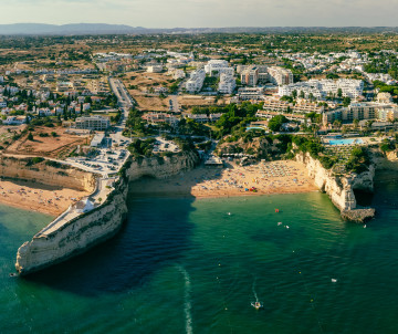 Aerial view of Praia do Carvoeiro in the Algarve, showing the sheltered sandy cove, turquoise Atlantic water, limestone cliffs and whitewashed buildings of the coastal village.