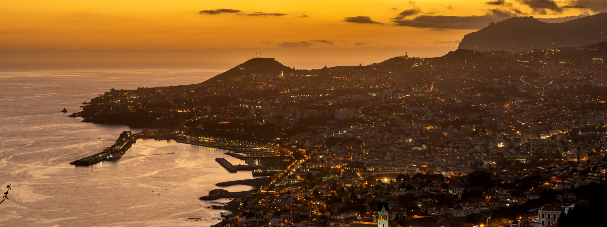 Panoramic view of Funchal, Madeira, Portugal, at sunset, featuring the city's waterfront, marina, and hillside neighborhoods illuminated by evening lights. The image captures the capital of Madeira overlooking the Atlantic Ocean beneath a golden sky.
