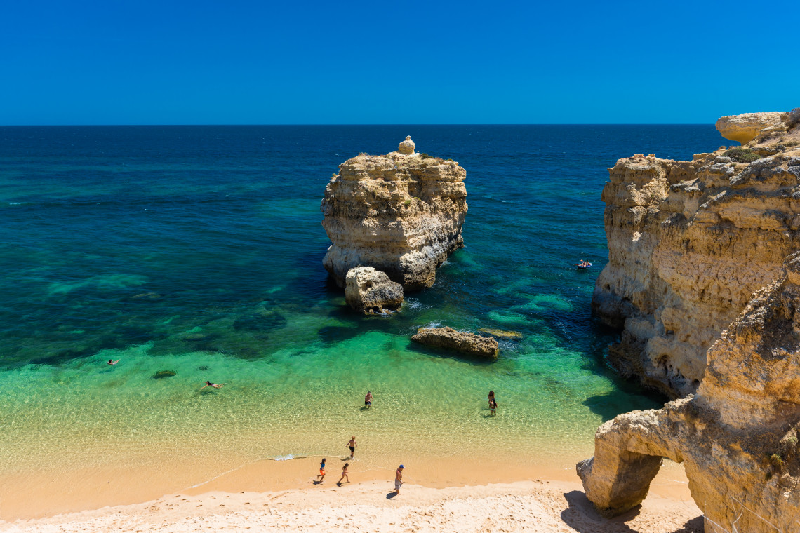 Golden sandy beach surrounded by limestone cliffs and turquoise Atlantic waters along the Algarve coast in Portugal. Visitors enjoy the sheltered cove, known for its dramatic rock formations, sea arches, and clear waters.