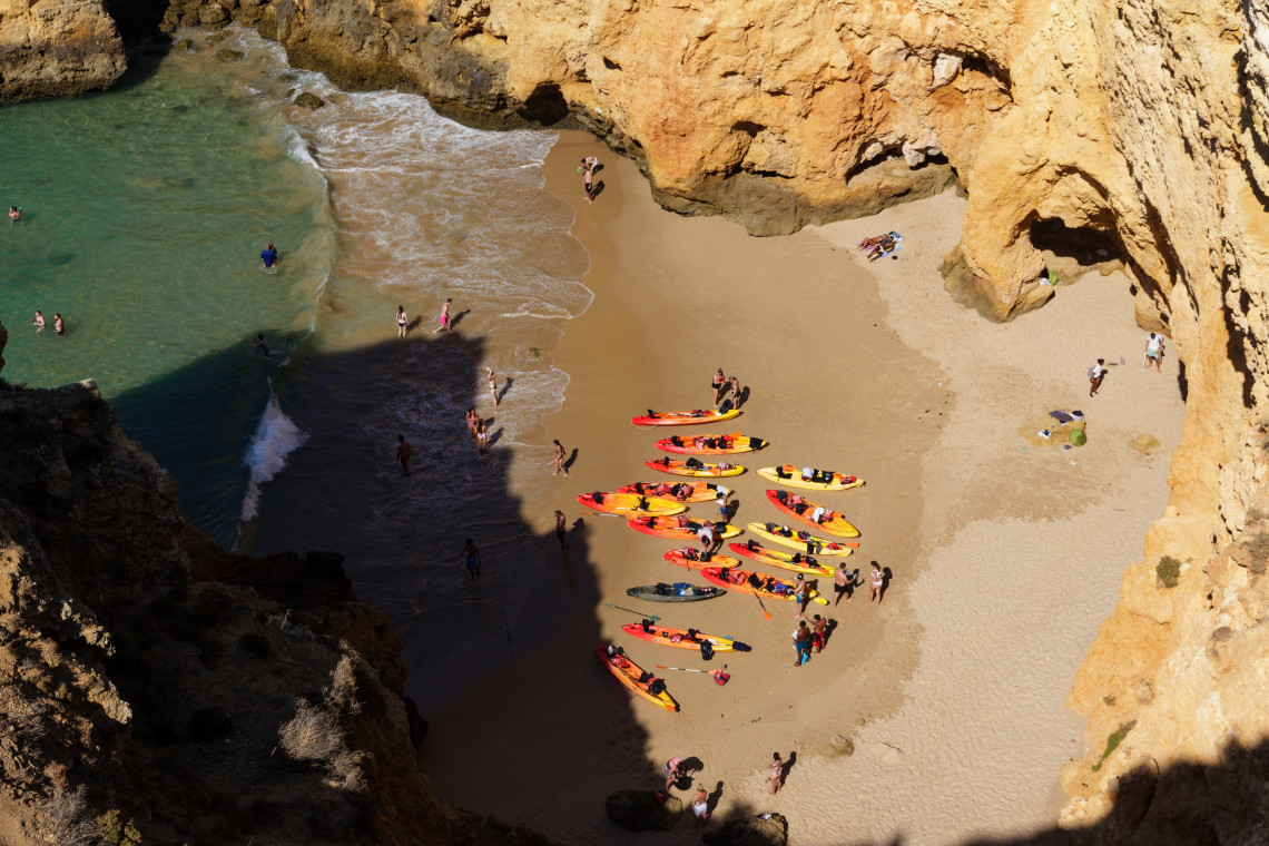 Aerial view of secluded beach surrounded by dramatic golden cliffs with multiple kayaks lined on sand, swimmers in turquoise waters, and visitors exploring the hidden cove in Algarve, Portugal