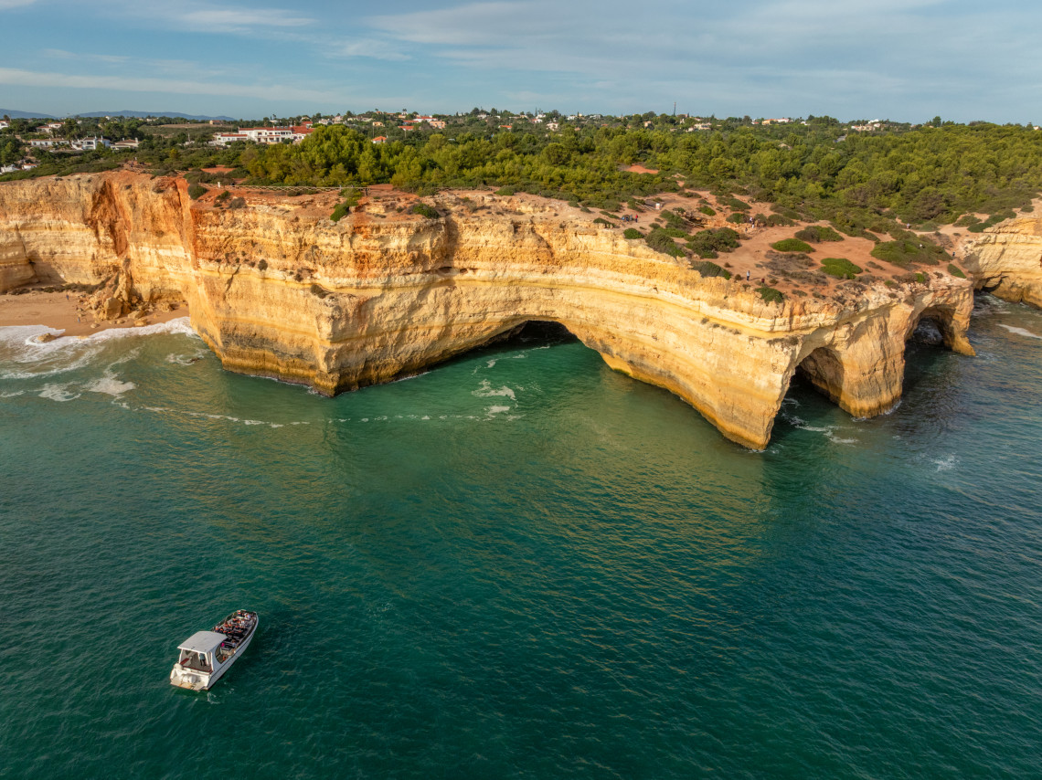 Aerial view of dramatic golden sandstone cliffs with natural sea arches and caves, turquoise Atlantic waters, small fishing boat, and Algarve coastline with vegetation and buildings in background, Portugal