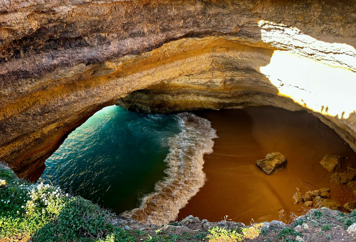 Natural sandstone rock arch formation framing turquoise ocean waters and white wave foam, sandy cove visible below golden stone layers, lush vegetation, dramatic geological feature on Portuguese coast