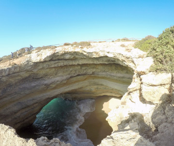 View from above of Benagil Sea Cave in the Algarve, Portugal, showing the natural limestone cave opening, sandy beach, and turquoise Atlantic waters entering through the sea arch. The image highlights one of the Algarve's most recognizable coastal rock fo