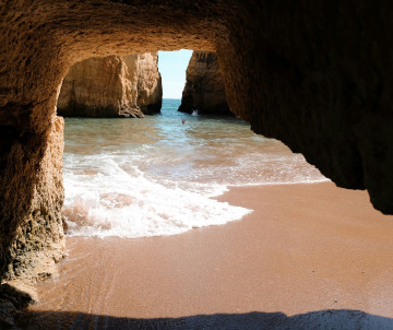 View from inside a golden limestone sea cave on the Lagoa coast, with a sandy cove, Atlantic waves and natural rock openings leading toward the ocean.