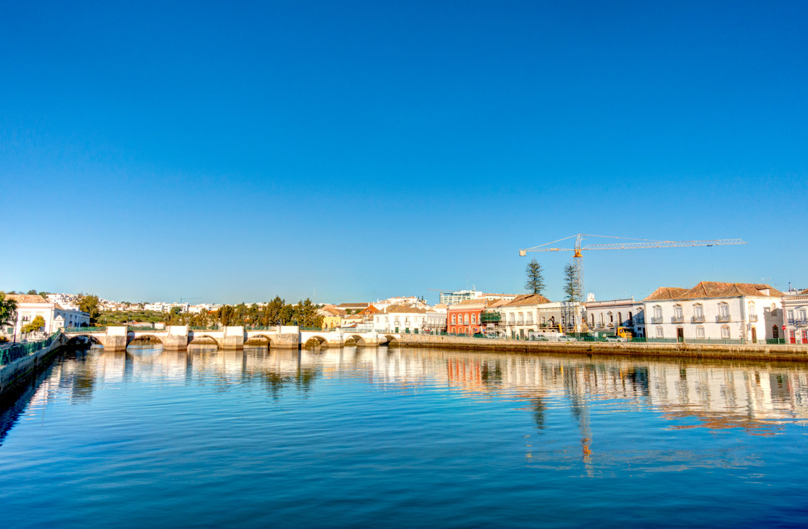View across the calm Gil&atilde;o River in Tavira, with traditional whitewashed buildings, waterfront architecture and the historic bridge reflected in the water.
