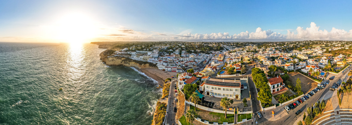 Panoramic aerial view of Carvoeiro at sunset, with whitewashed houses and red-tiled roofs clustered above a sandy Atlantic cove and golden limestone cliffs.