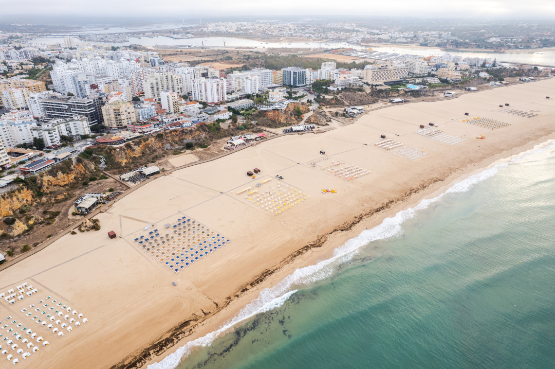 Aerial view of Praia de Alvor near Portim&atilde;o, showing its expansive sandy beach, beachfront buildings and the lagoon and wetlands of the Alvor Estuary behind the coast.