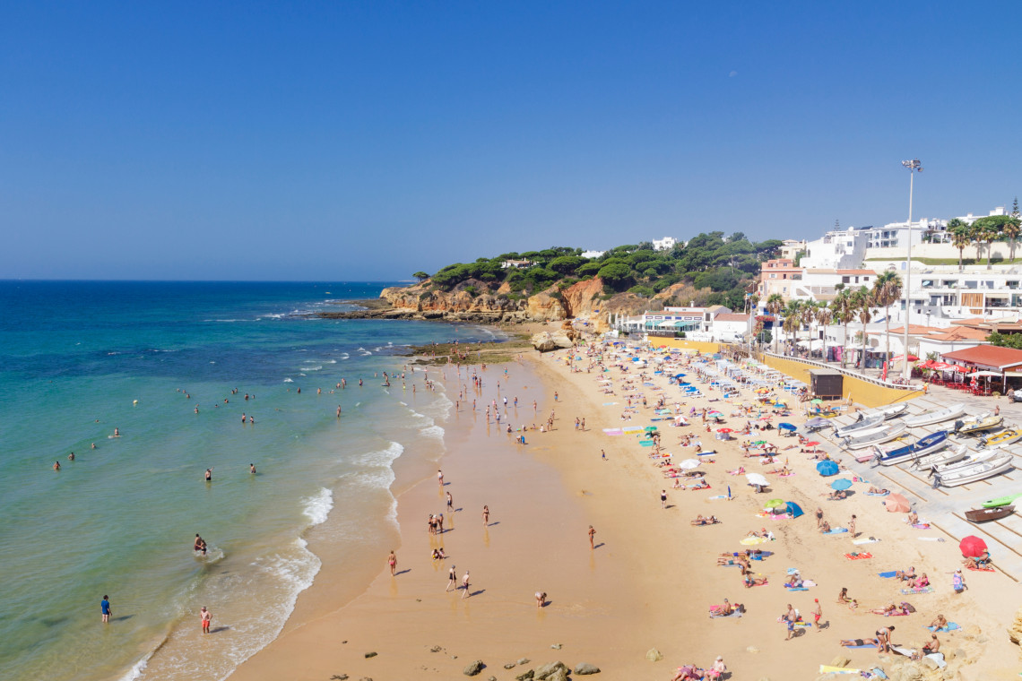 A busy sandy beach on the Algarve coast with swimmers, sun umbrellas and waterfront buildings beside calm Atlantic waters.