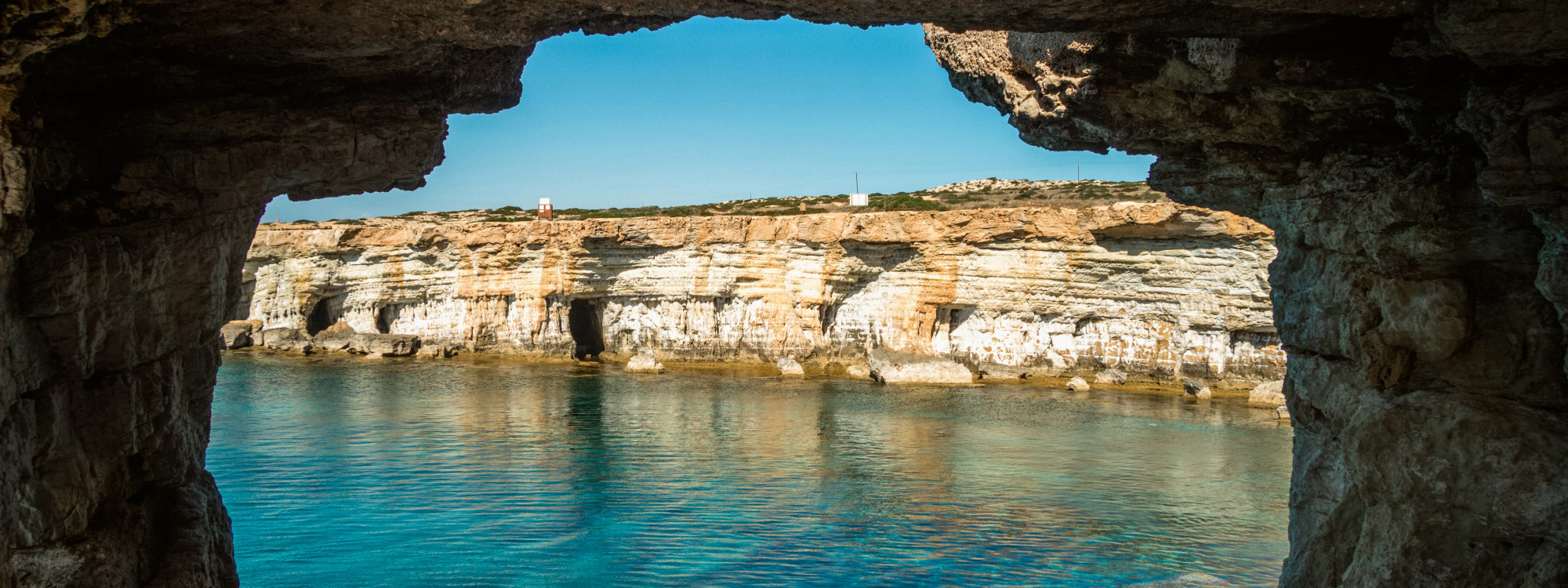 View from inside a coastal sea cave near Lagoa in the Algarve, Portugal, looking across clear blue water toward layered limestone cliffs. The dark cave walls naturally frame the sunlit coastline.