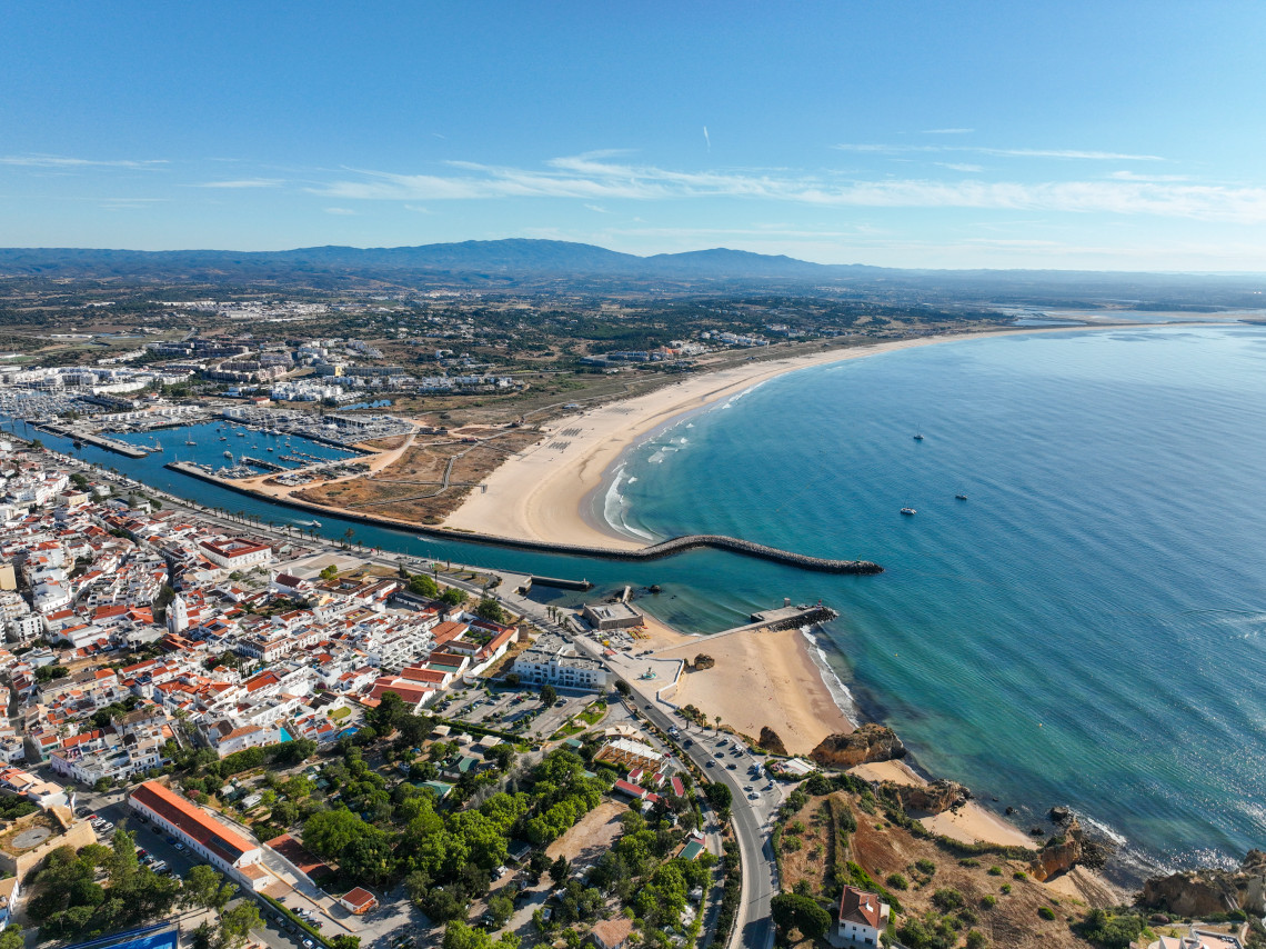 Aerial view of Lagos in the Algarve, Portugal, showing the marina, harbour entrance and the long sandy shoreline of Meia Praia. White buildings, breakwaters and calm Atlantic waters extend across the coastal landscape.