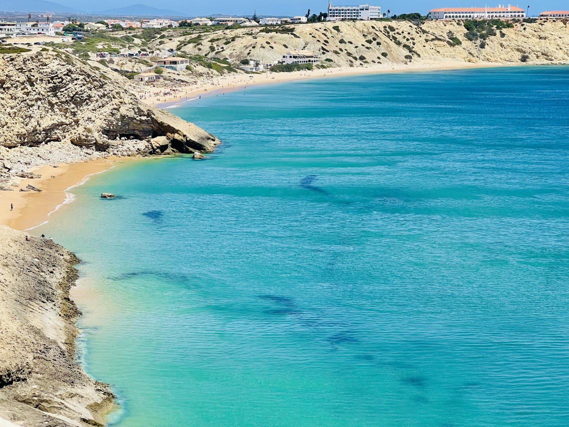 Elevated view of Praia do Porto de M&oacute;s in Lagos, Algarve, Portugal, with a wide sandy beach curving around clear Atlantic water. Pale limestone cliffs and buildings rise behind the shoreline.