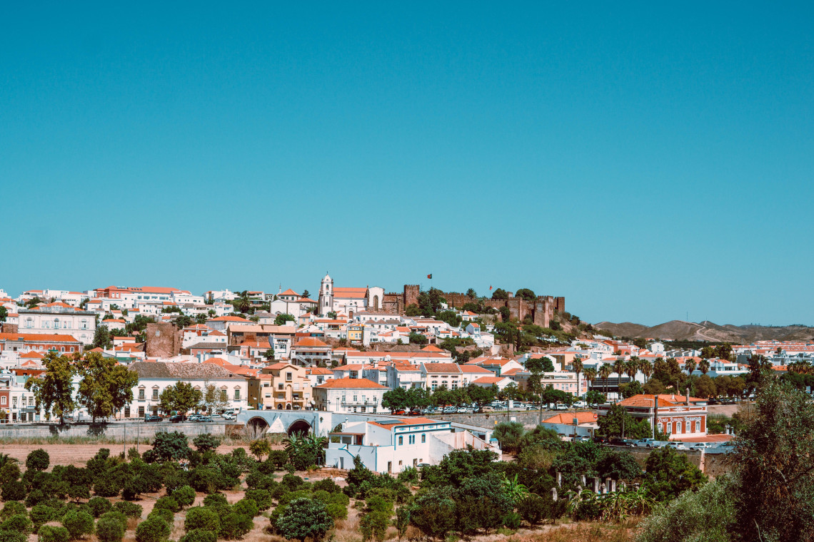 Panoramic view of Silves in the Algarve, Portugal, with whitewashed houses and terracotta roofs surrounding the hilltop castle. Trees, orchards and historic buildings fill the foreground.