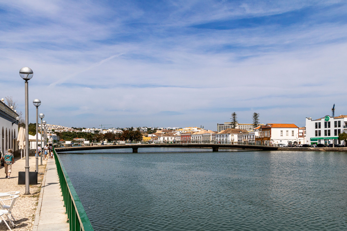 The Gil&atilde;o River waterfront in Tavira, Algarve, Portugal, viewed from a paved promenade with green railings. A bridge crosses the calm river between rows of whitewashed buildings.