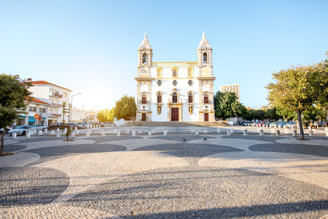 Front view of Igreja do Carmo in Faro, Algarve, Portugal, featuring an ornate white-and-yellow fa&ccedil;ade and two bell towers. The church faces a spacious square paved with patterned black-and-white Portuguese cobblestones.