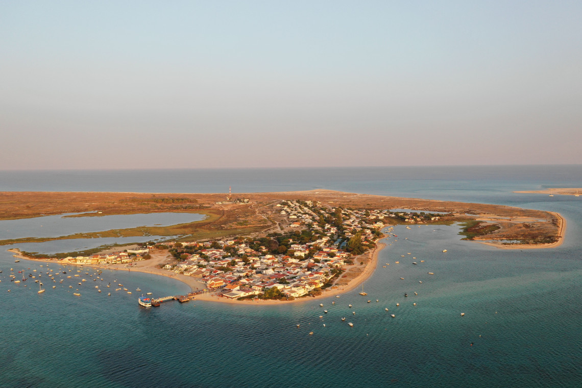 Aerial view of Ilha da Culatra in Faro, Algarve, Portugal, showing a fishing village surrounded by the channels and sandbanks of the Ria Formosa. Small boats are anchored near the houses, with dunes and the Atlantic Ocean beyond.