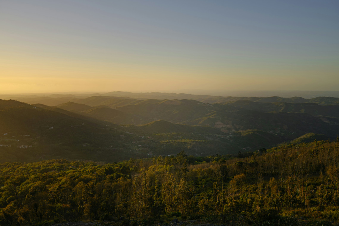 Layered hills and forested valleys in the Serra de Monchique, Algarve, Portugal, illuminated by low evening sunlight. The mountainous landscape extends toward the hazy horizon.