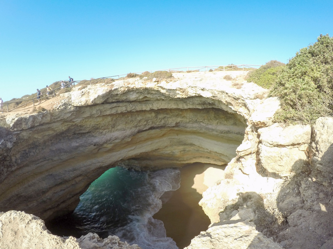 A limestone sea cave at Algar Seco in Carvoeiro, Algarve, Portugal, viewed from the cliff above. Atlantic waves enter the eroded rock opening beneath a fenced coastal footpath.