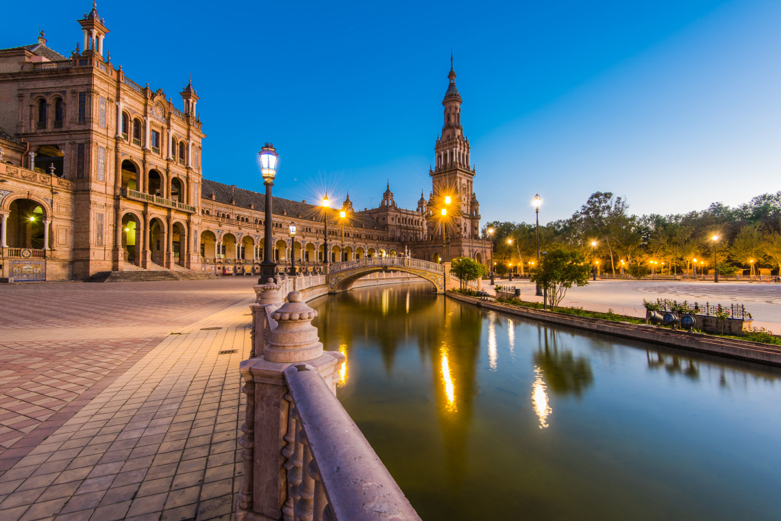 Plaza de Espa&ntilde;a in Seville, Spain, illuminated at dusk beside its curved canal. Ornate arcades, a tiled bridge and the central tower are reflected in the still water.