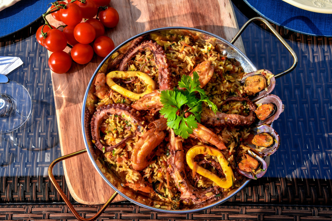 Traditional seafood rice served in the Algarve, Portugal, topped with grilled octopus, prawns, mussels and squid rings. The dish is presented in a large metal pan with fresh parsley and tomatoes alongside it.