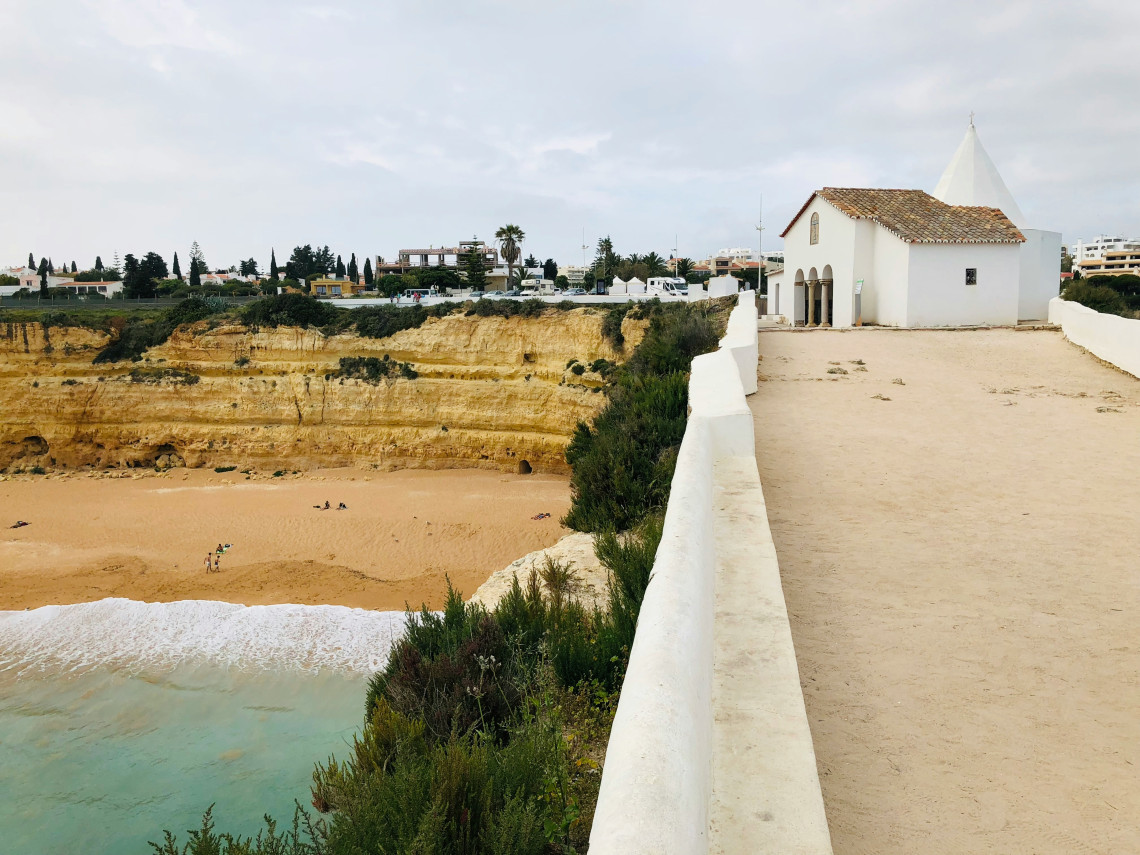 The whitewashed Chapel of Nossa Senhora da Rocha stands on a narrow clifftop promontory in Porches, overlooking a sandy beach and the Atlantic Ocean.