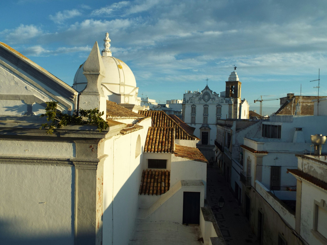 View across the whitewashed rooftops and traditional terracotta tiles of Faro&rsquo;s historic center, with ornate church towers rising above the narrow streets.