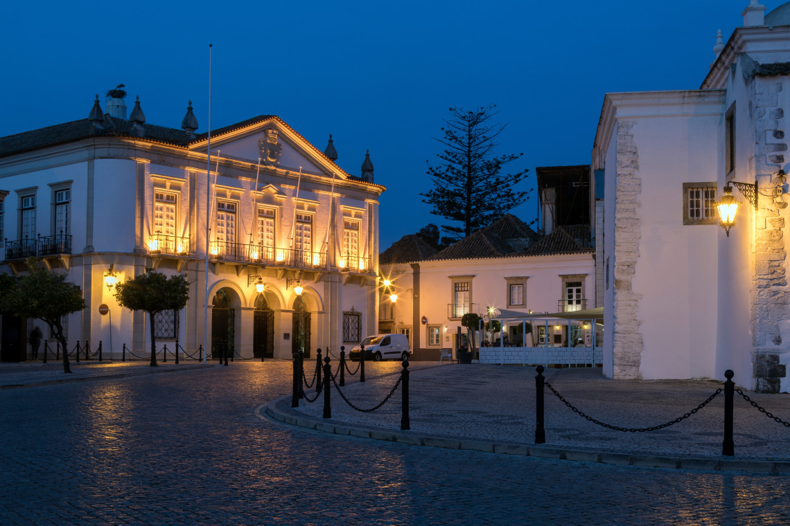 Illuminated historic buildings surround a cobbled square in Faro at dusk, with traditional Algarve architecture, street lamps and mature trees creating an atmospheric evening scene.