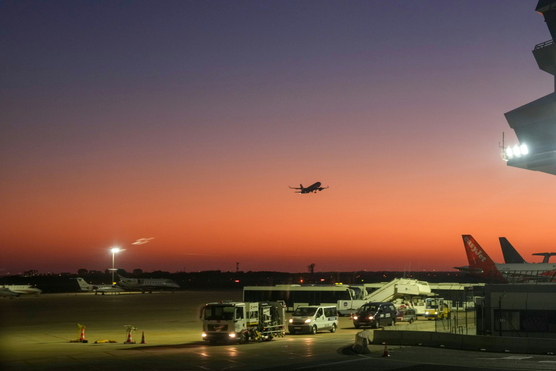 An aircraft takes off from Faro Airport in Faro, Algarve, Portugal, against an orange sunset sky. Ground service vehicles and parked planes are visible across the airport apron.