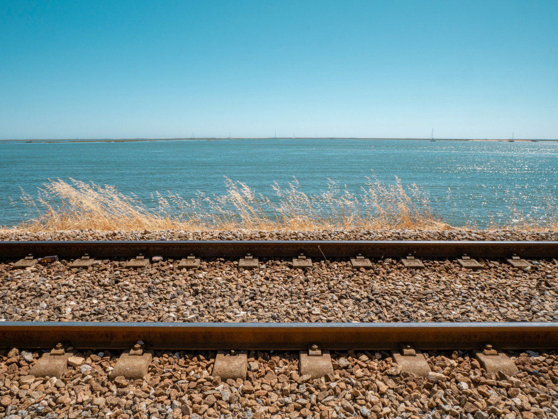Railway tracks run alongside the blue waters and salt-marsh landscape of Ria Formosa near Faro in the Algarve, Portugal. The open lagoon and distant sailboats reflect the region&rsquo;s distinctive coastal setting.