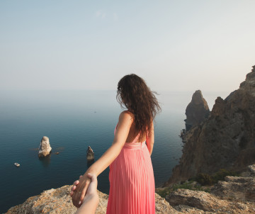 A traveler stands on the cliffs of Ponta da Piedade in Lagos, Algarve, Portugal, overlooking the Atlantic Ocean while holding a companion's hand. The viewpoint features the region's distinctive limestone rock formations rising from the sea.