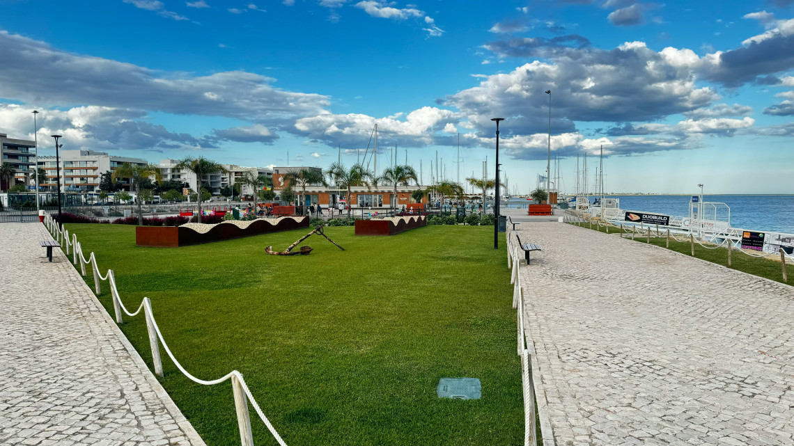 A landscaped waterfront promenade with green lawns, paved walkways and sailboat masts overlooks a marina on Portugal&rsquo;s Algarve coast.